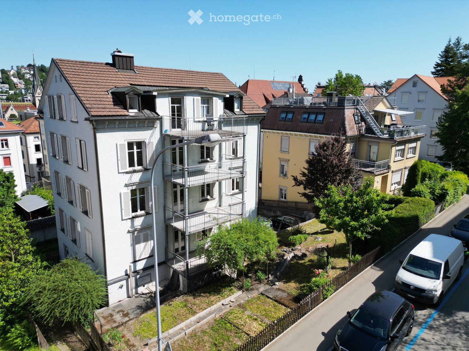 Multi-story residential building with balconies, surrounded by greenery and parked cars.