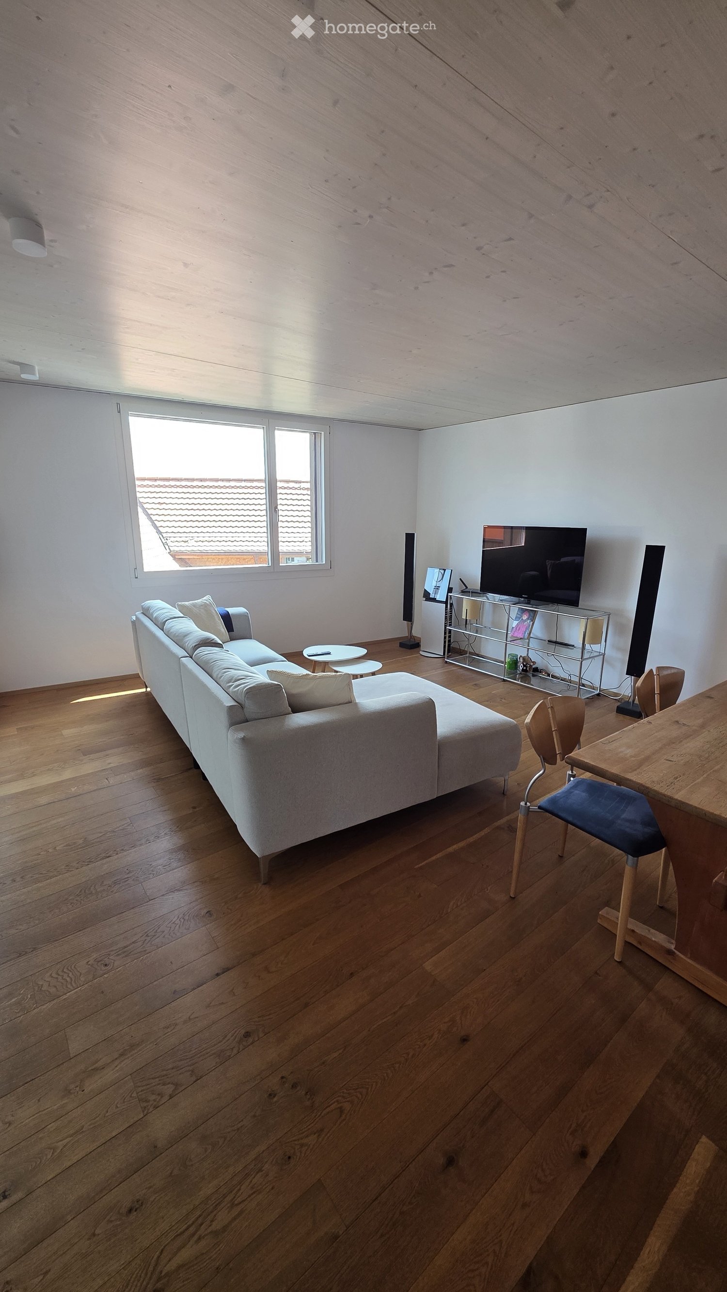Modern living room with light wooden flooring, white walls, and large windows. Features a grey sectional sofa, small round tables, a wooden dining table with chairs, and a television on a metal stand.