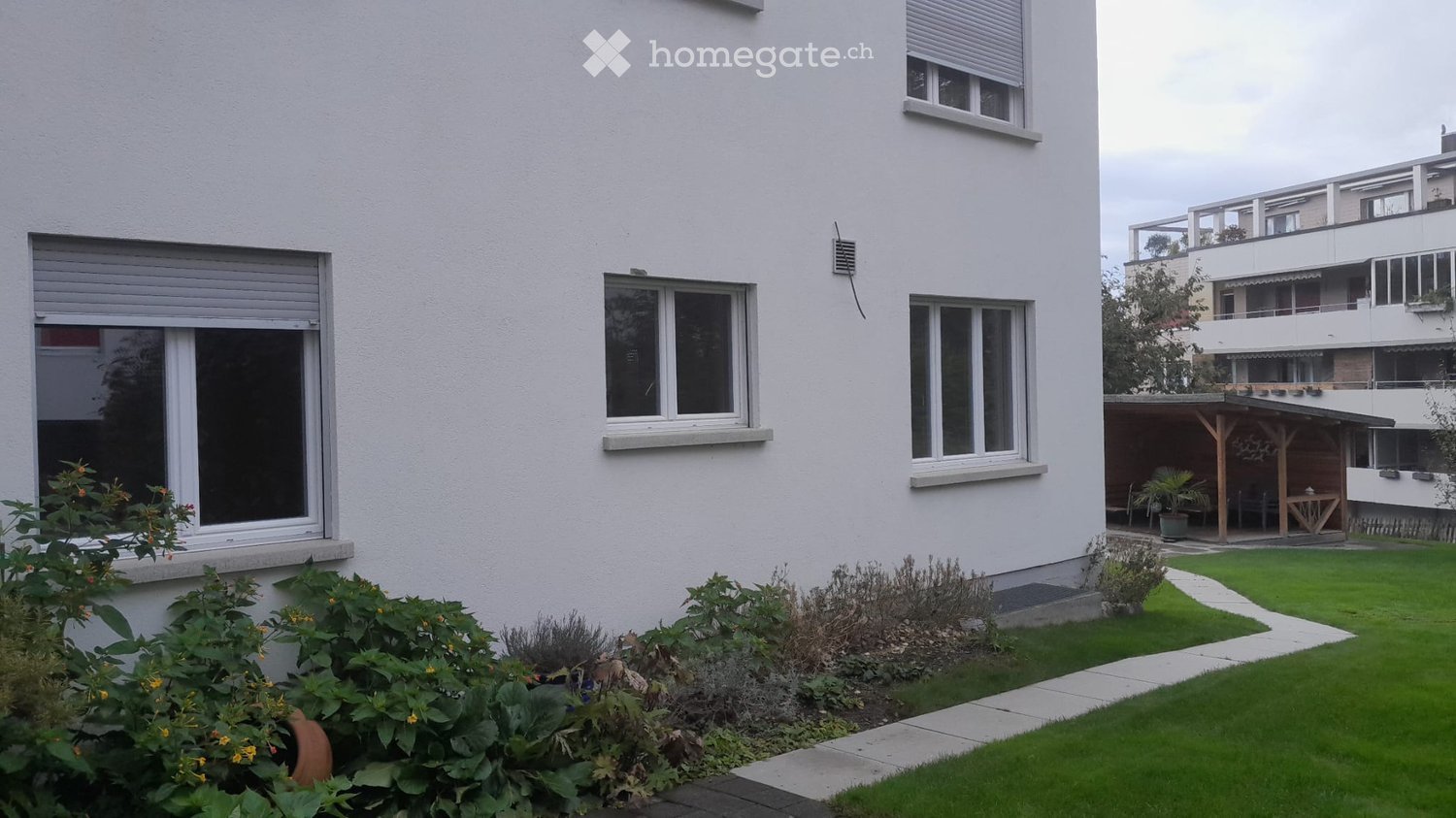 Modern white building with multiple windows, shutters, and a well-maintained garden with green lawn, shrubs, and flowers.