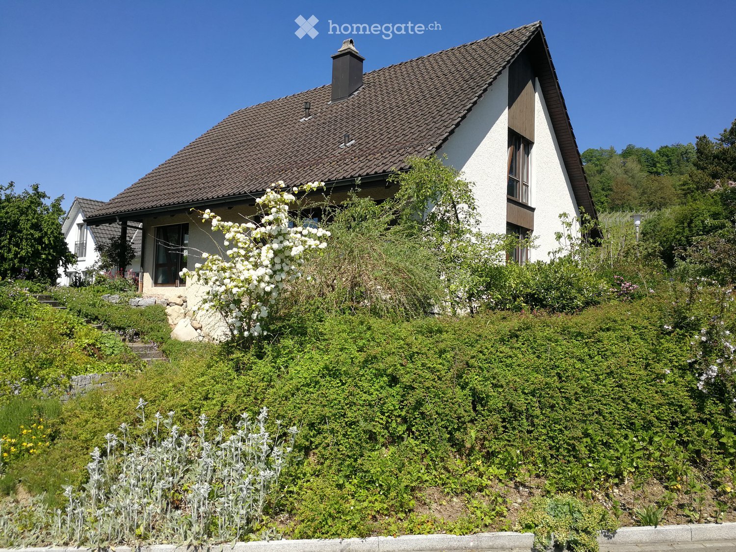 Two-story house with a tiled roof, white walls, and a chimney, surrounded by a well-maintained garden with flowering shrubs and trees.