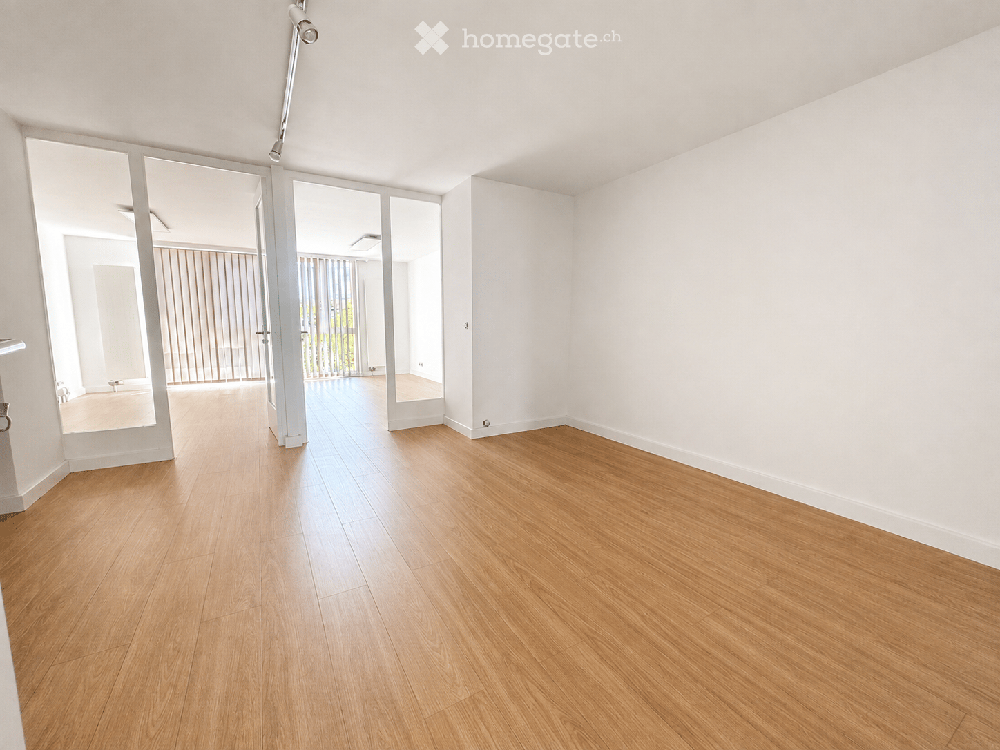 Empty room with light wooden flooring and white walls, featuring large sliding glass doors leading to another space.