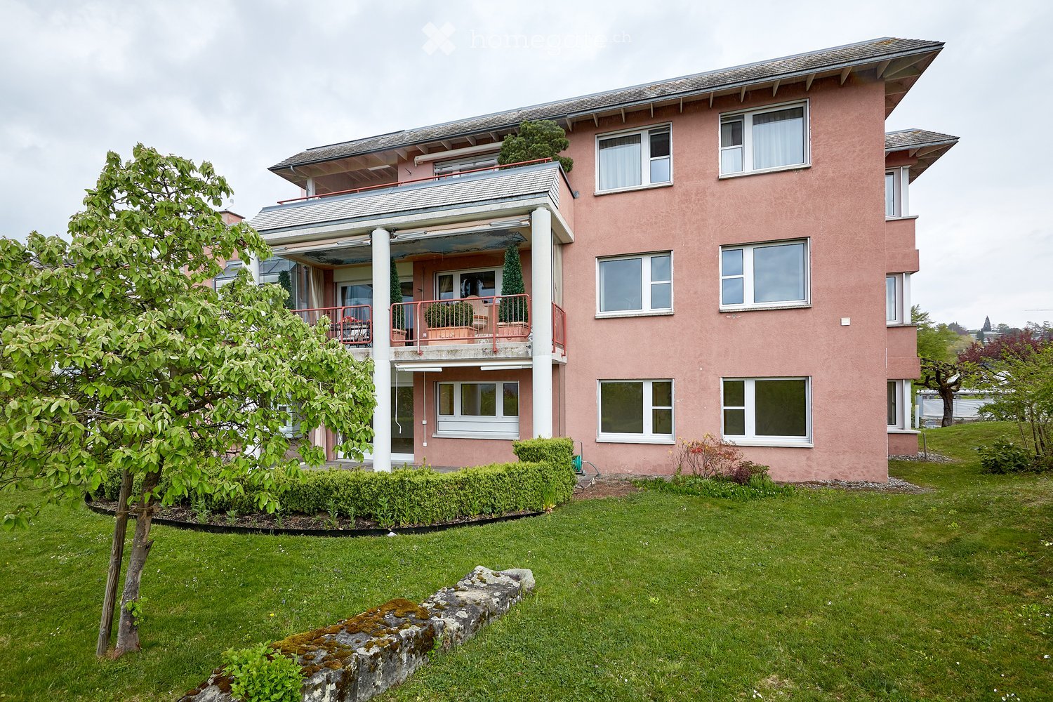 Pink multi-story building with white-framed windows, a balcony, and surrounding greenery including a lawn, shrubs, and trees.