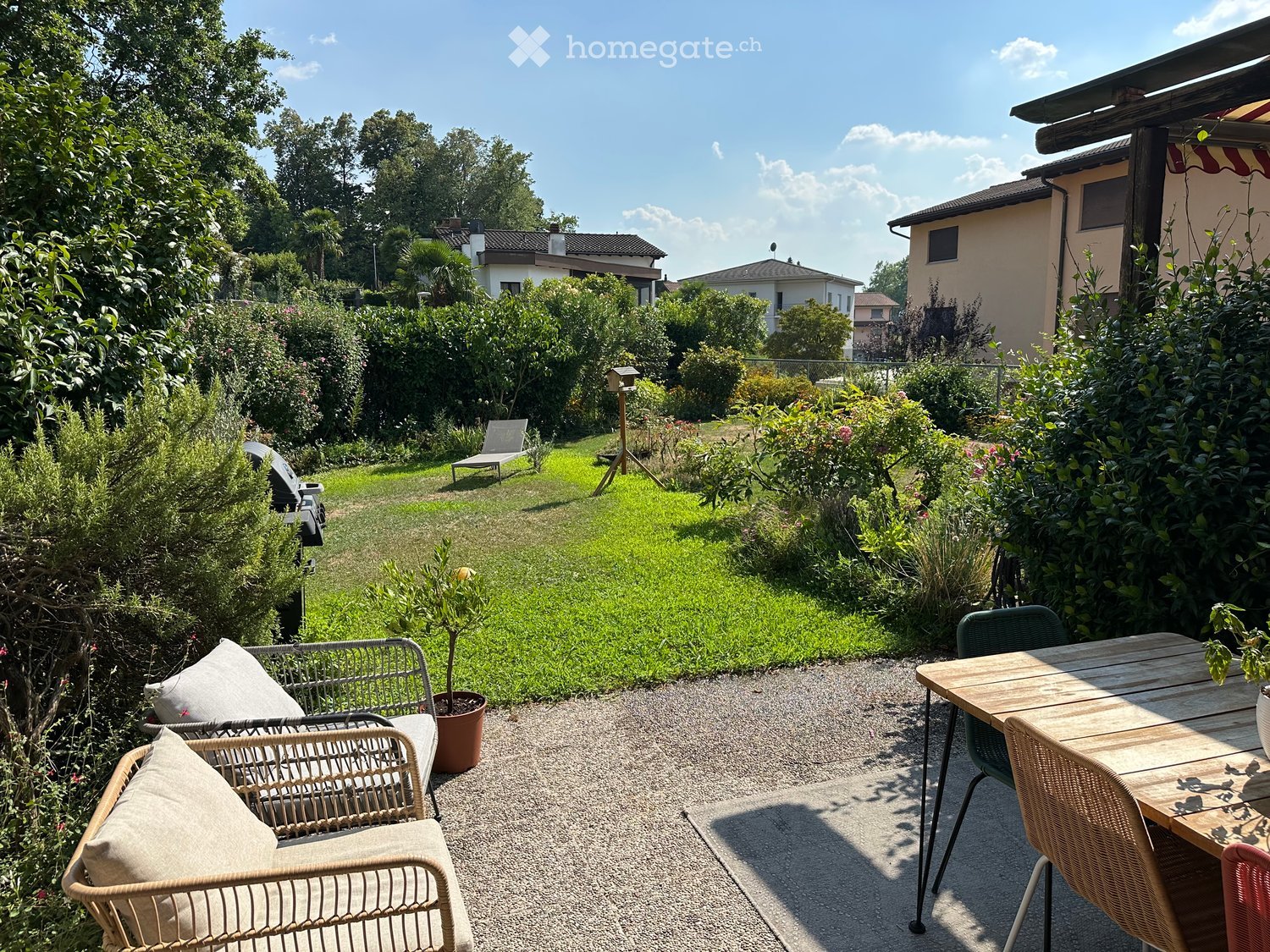 Well-maintained garden with a gravel patio area featuring a wooden table, wicker chairs, and a lounge chair. The garden includes lush greenery, a grassy lawn, and a small potted plant.