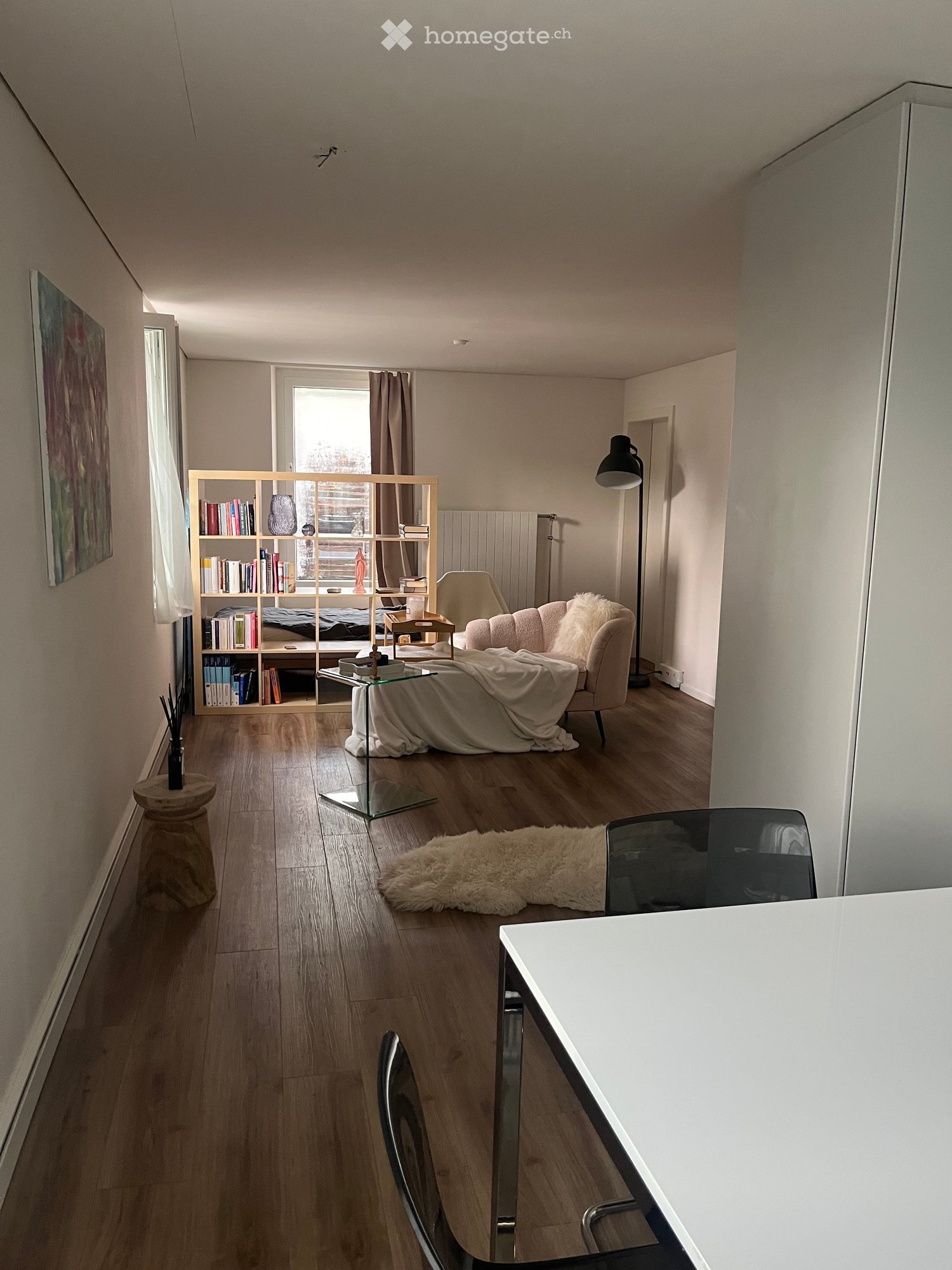 Modern living room with wooden flooring, a cream-colored sofa, a glass coffee table, a bookshelf, a floor lamp, and a white desk with chairs.