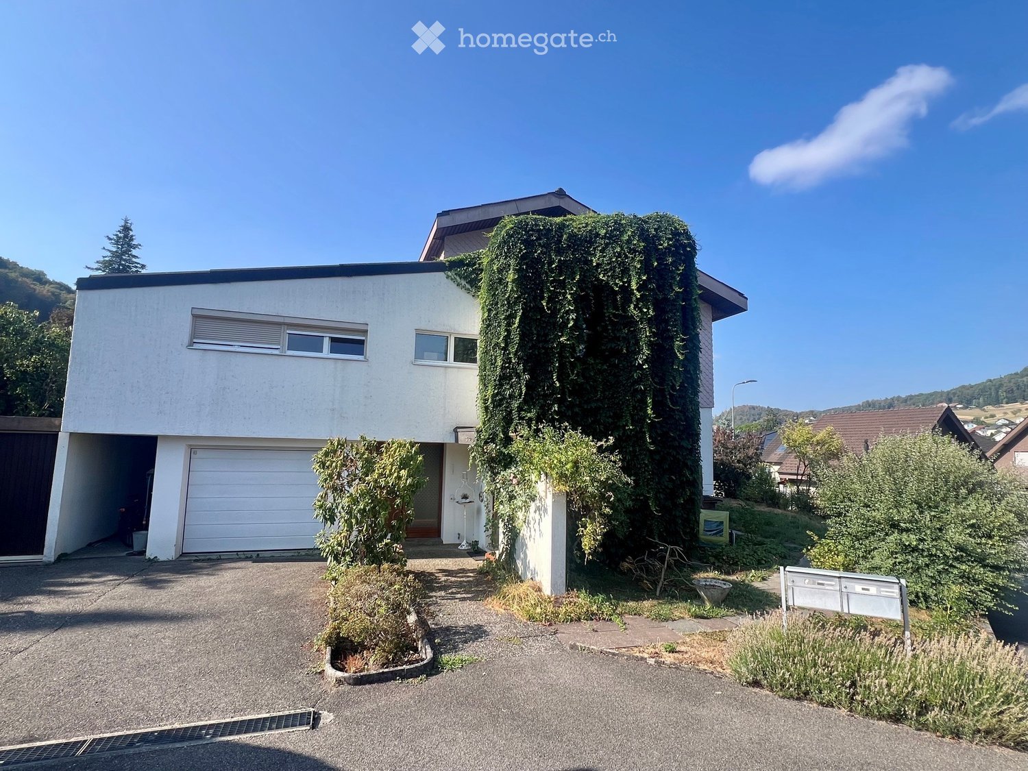 Two-story house with white exterior, garage, and ivy-covered wall. The house features a driveway and is surrounded by greenery and a clear blue sky.