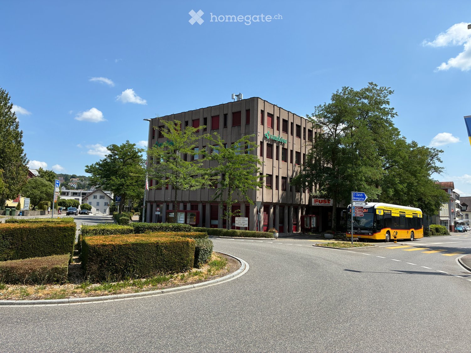A multi-story building with a modern facade, surrounded by greenery and a road with a bus passing by.