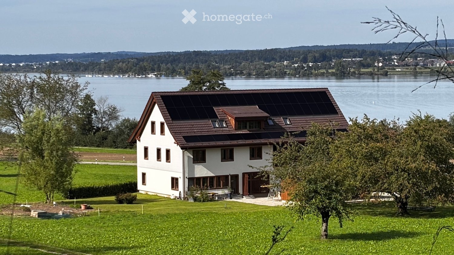 A two-story white house with a brown roof and solar panels, situated on a grassy lawn near a lake with trees and distant hills.