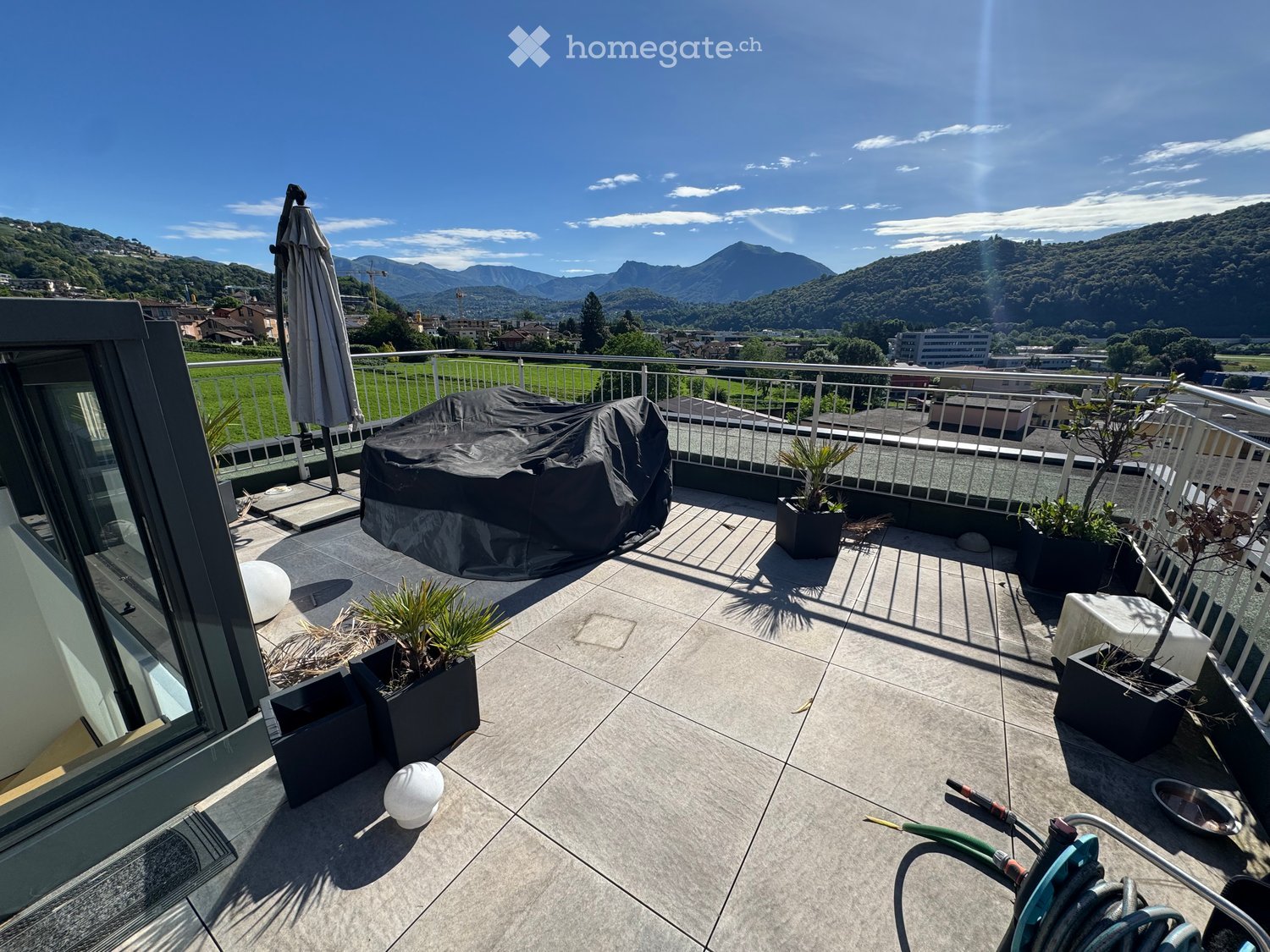 Balcony with tiled floor, metal railing, potted plants, covered furniture, and scenic mountain view.