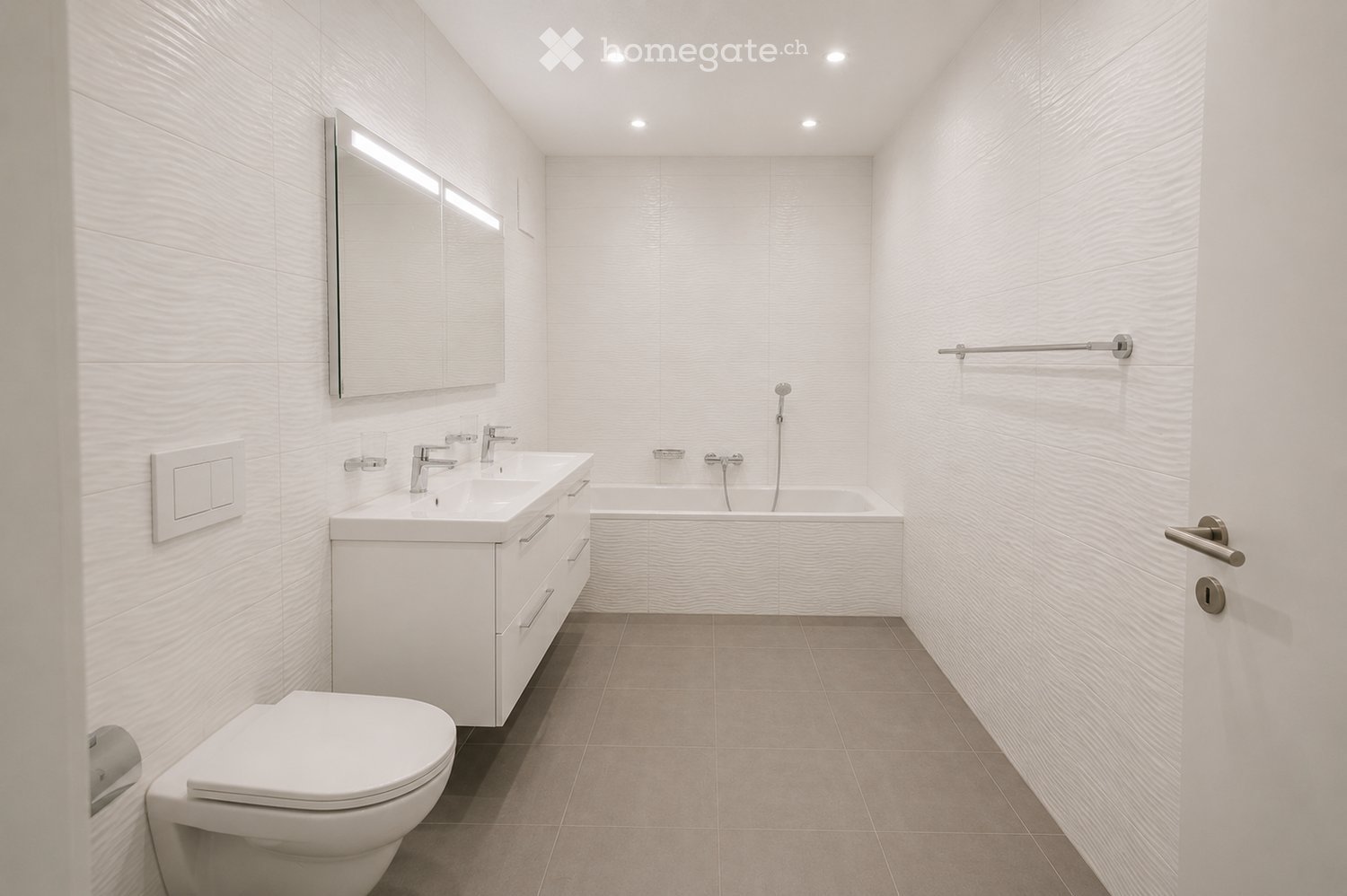 Modern bathroom with white tiles, a double sink vanity, a bathtub, a toilet, and a large mirror with built-in lighting.