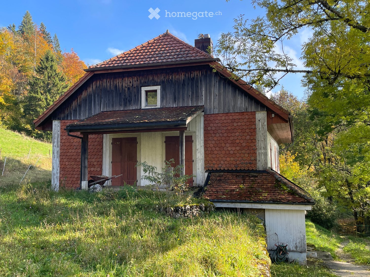 A rustic wooden house with a red-tiled roof, surrounded by trees and greenery. The house features a small porch with a white door and red shutters, and a small garden area in front.