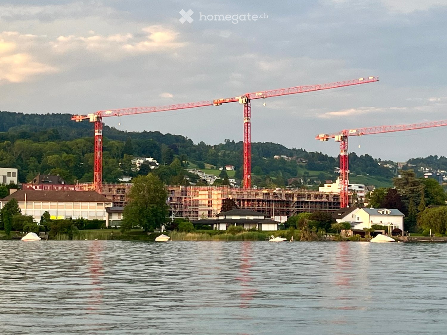 A construction site with two large cranes and scaffolding, situated near a body of water with surrounding greenery and existing buildings.