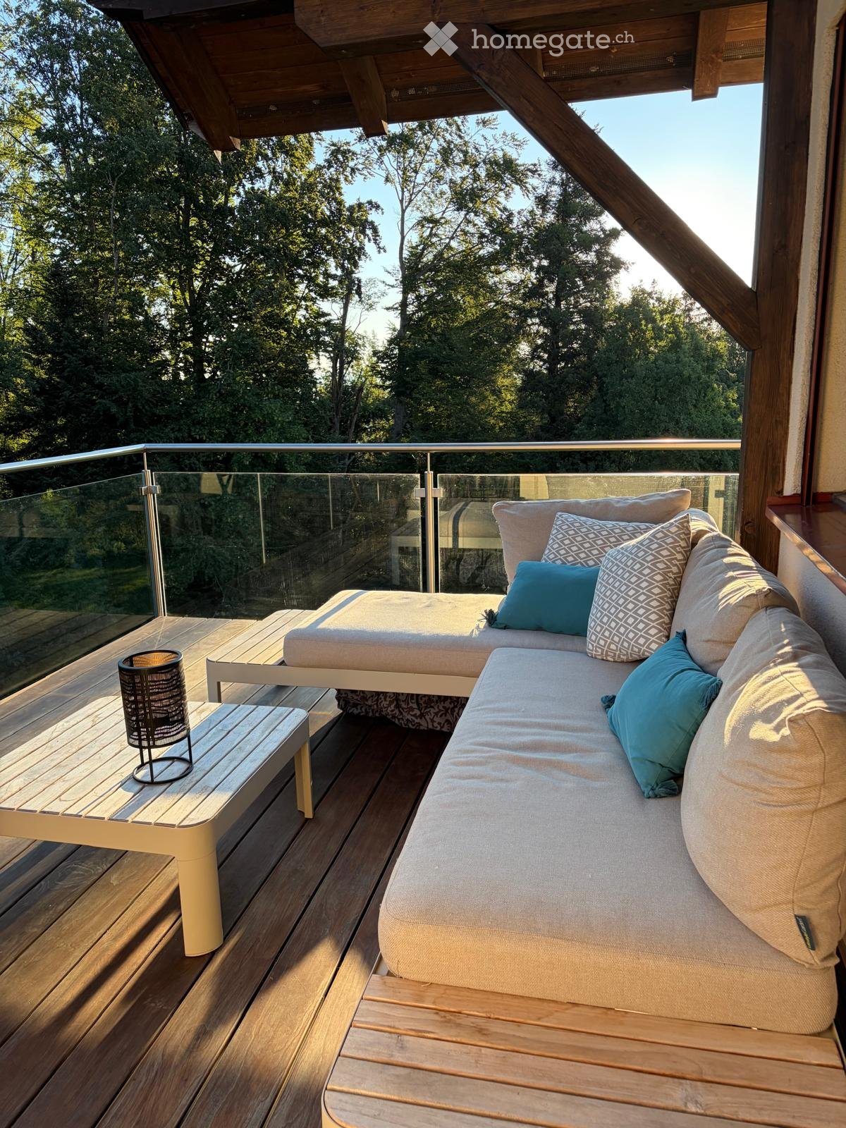A modern wooden deck with a glass railing, featuring a beige sofa with blue and patterned cushions, a small wooden bench, and a decorative metal candle holder.