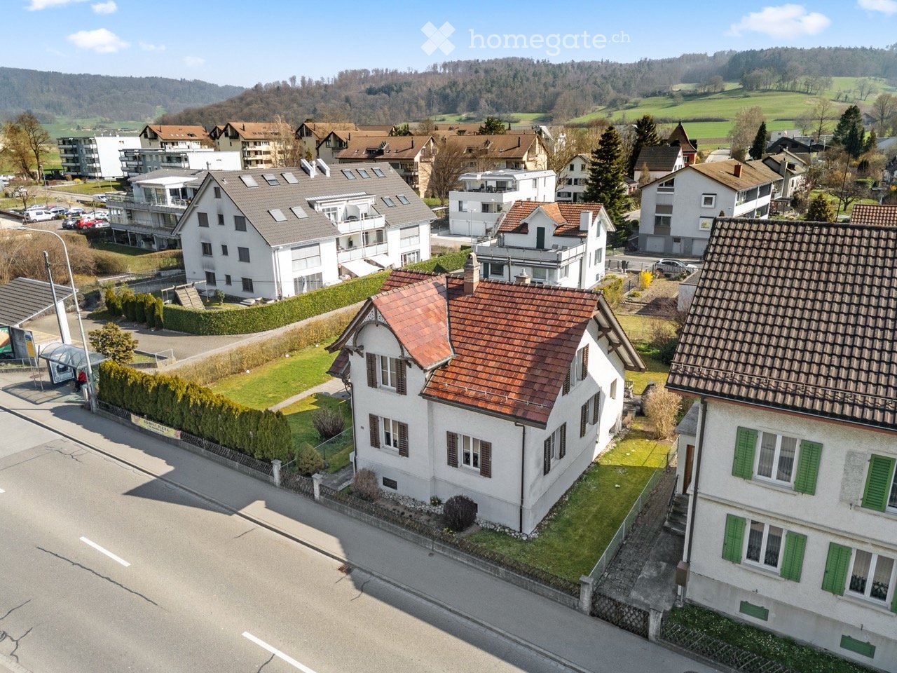 Aerial view of a residential area with multiple houses, green lawns, and a hilly landscape in the background.