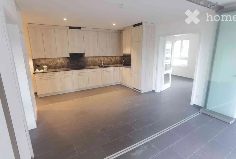 Modern kitchen with light wooden cabinets, stainless steel appliances, and a dark tiled floor. The space is open and bright with large windows.