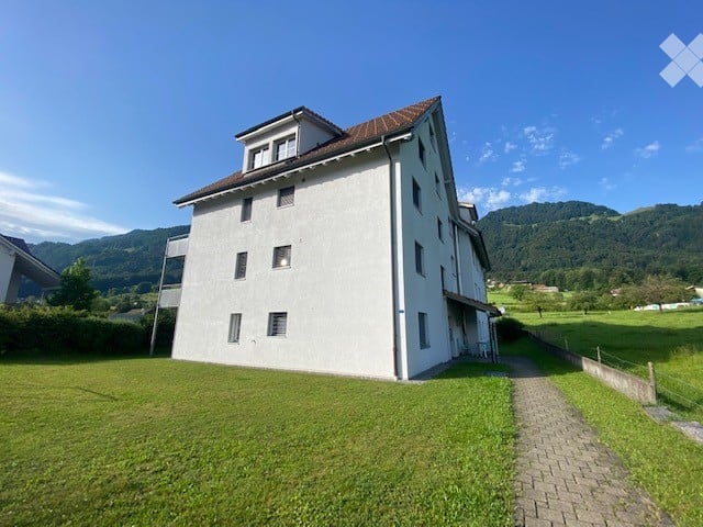 A modern, white, multi-story building with a brown roof, surrounded by a green lawn and a scenic mountain view in the background.