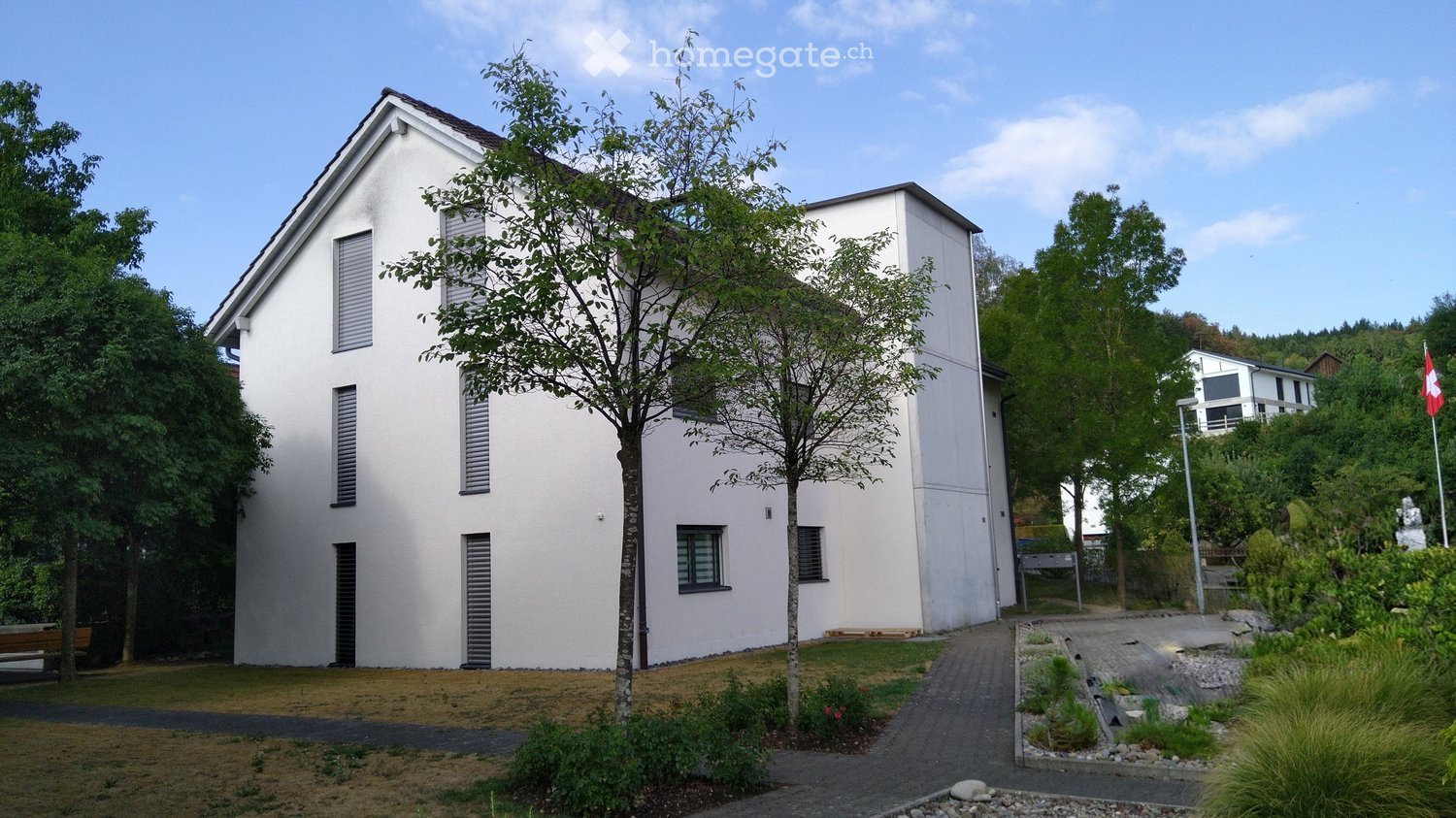 Modern two-story white house with large windows and shutters, surrounded by greenery and a paved pathway.