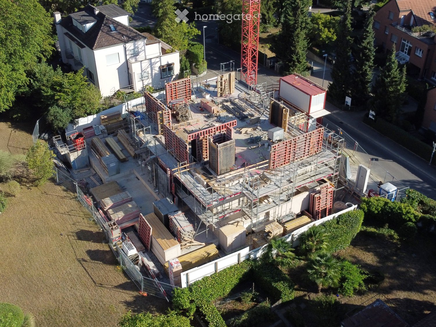 Aerial view of a construction site with scaffolding, building materials, and partially constructed structures. Surrounding area includes existing houses and greenery.