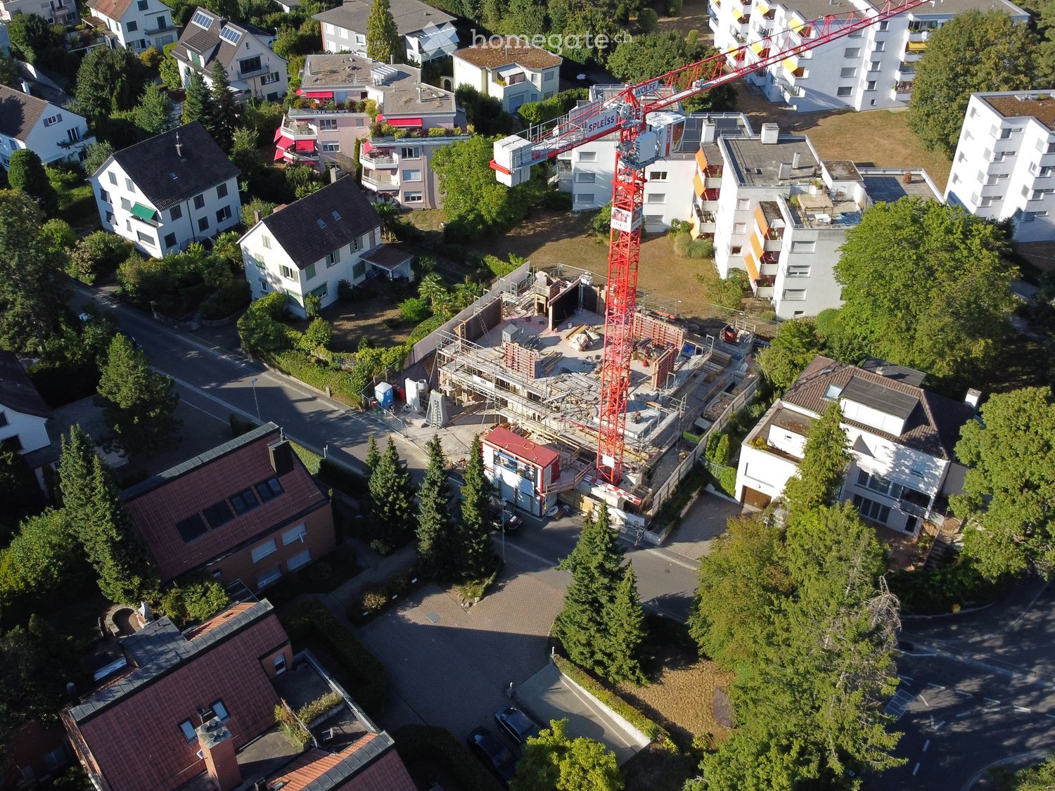 Aerial view of a residential area with multiple houses, a construction site with a crane, and surrounding greenery.