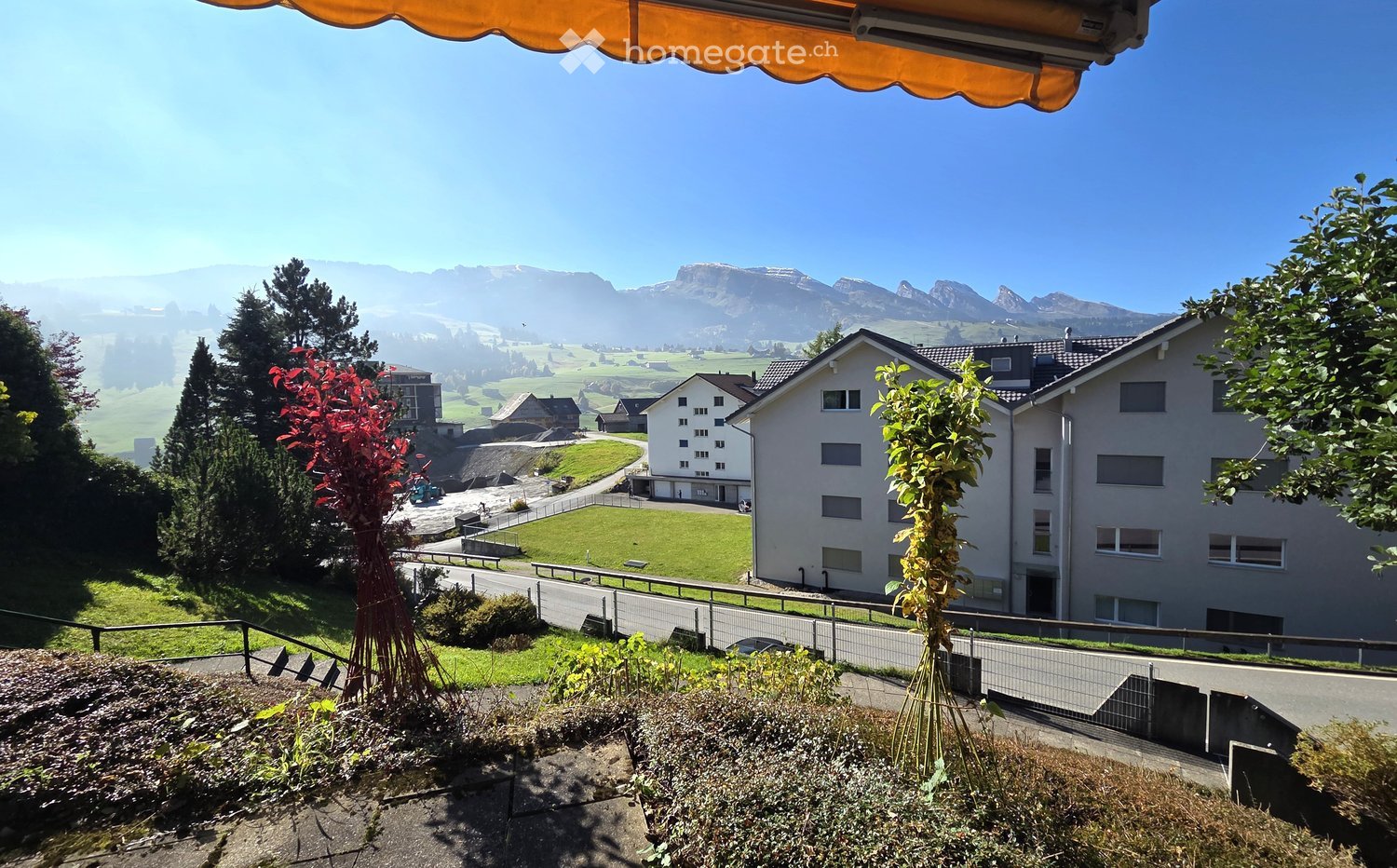 Balcony with an orange awning overlooking a scenic valley with houses, green fields, and distant mountains.