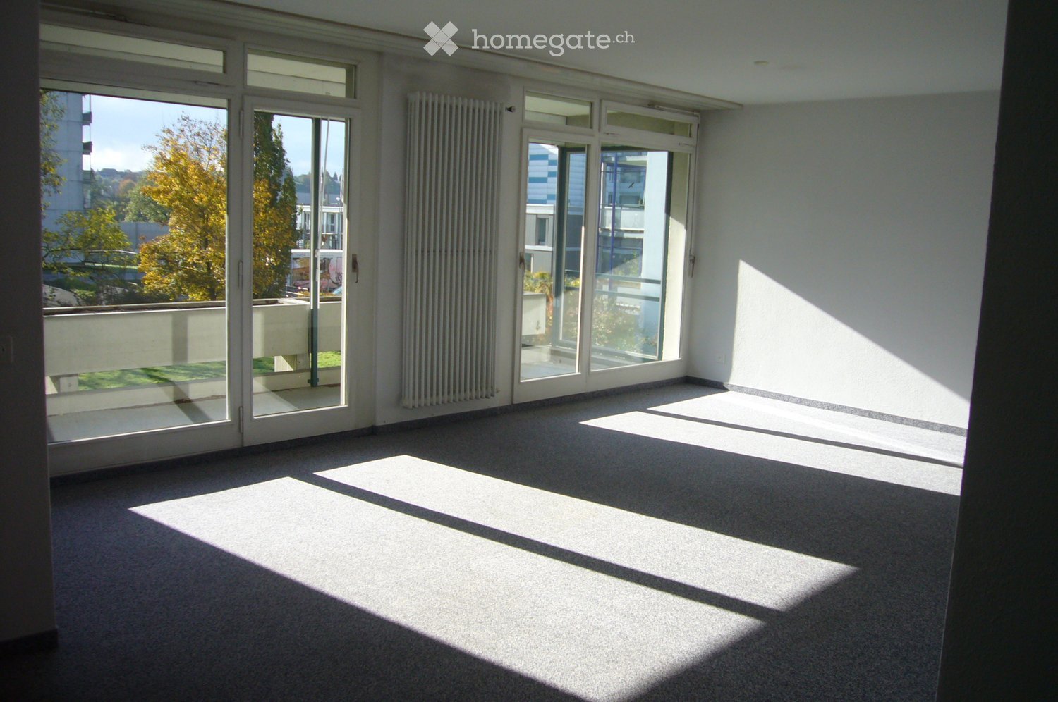 Empty room with large windows and sliding glass doors leading to a balcony, carpeted floor, radiator, and natural light streaming in.
