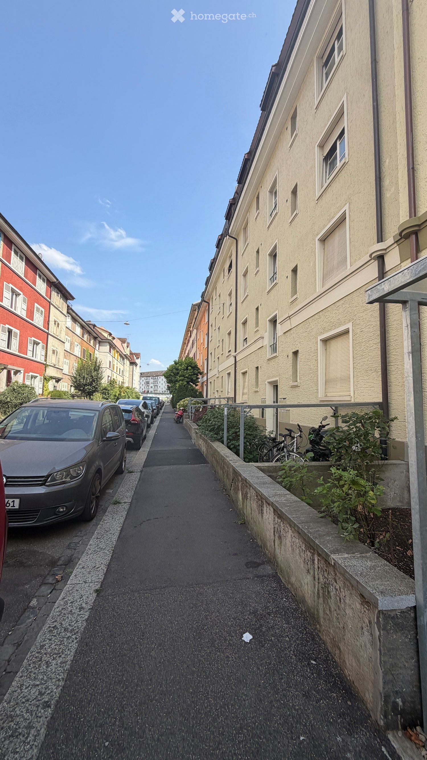 Narrow urban alleyway flanked by multi-story residential buildings, with parked cars and bicycles along the sides.