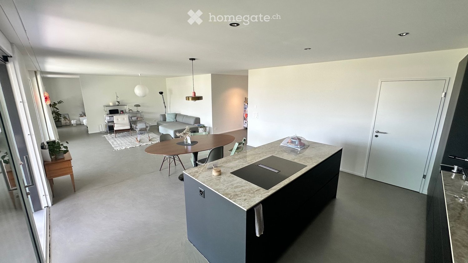 Modern open-plan living and dining area with a grey sofa, coffee table, dining table, chairs, and a kitchen island with a sink and faucet. The space features pendant lights, a plant, and a sleek, contemporary design.