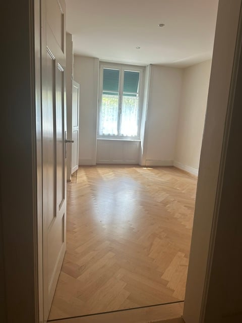Empty room with wooden herringbone flooring, white walls, and a window with shutters. No furniture or equipment present.