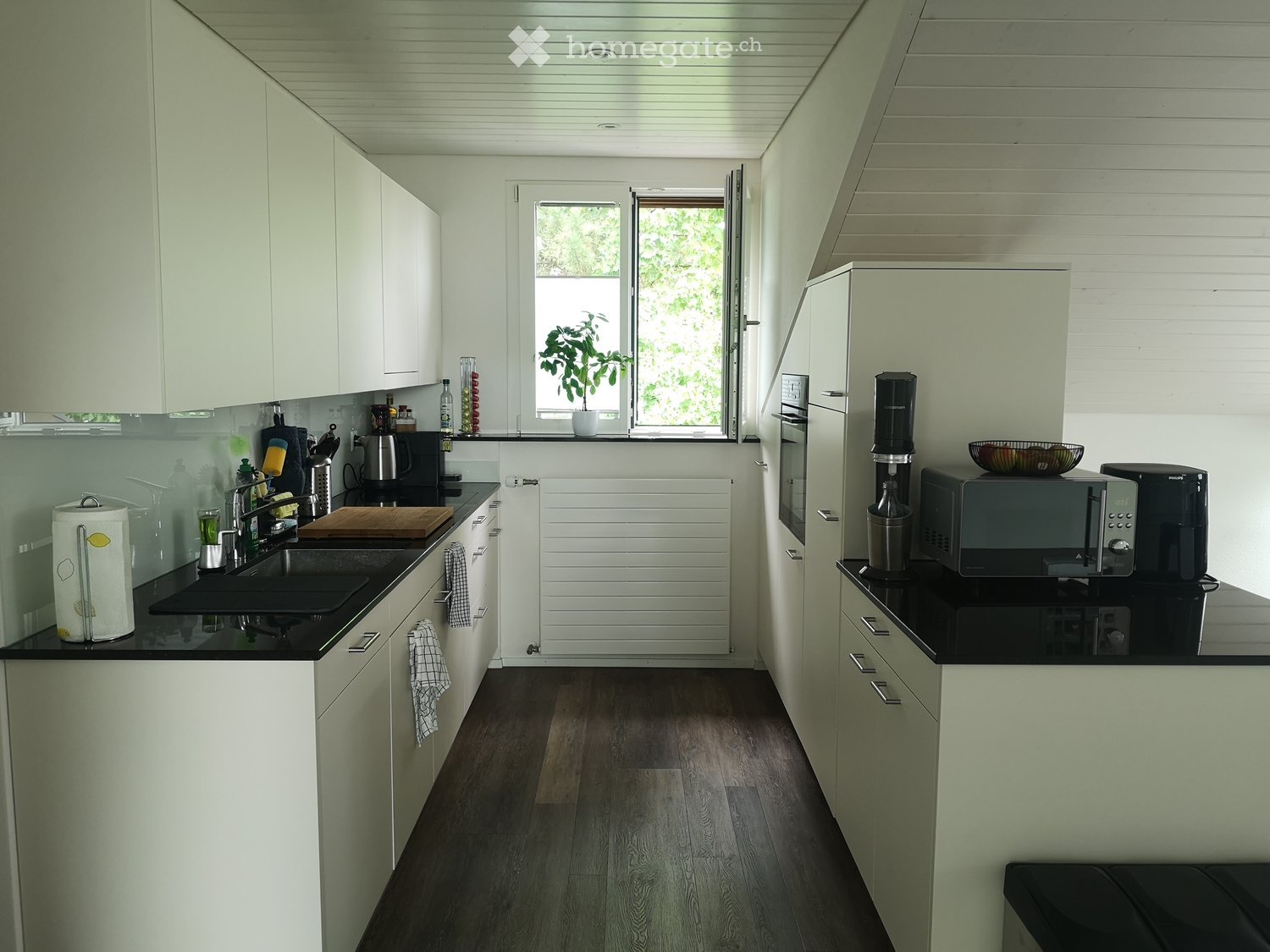 Modern kitchen with white cabinets, dark countertops, and various appliances including a microwave, coffee maker, and oven. There is a window with a plant on the sill and a sink with a faucet.