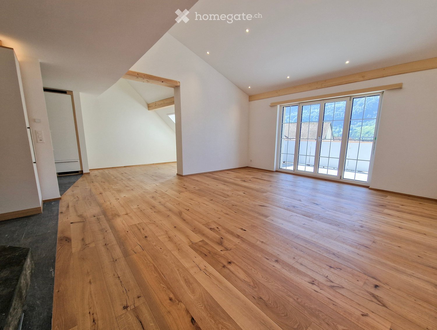 Empty room with wooden flooring, white walls, and large windows with a view of greenery outside.