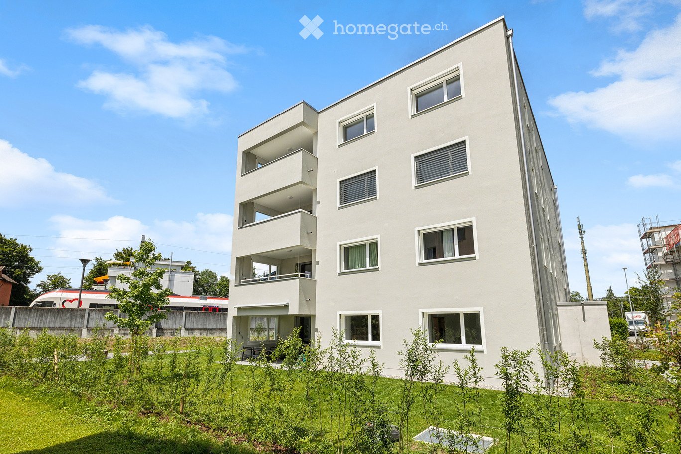 Modern, multi-story residential building with balconies and large windows, surrounded by greenery and a small garden area.