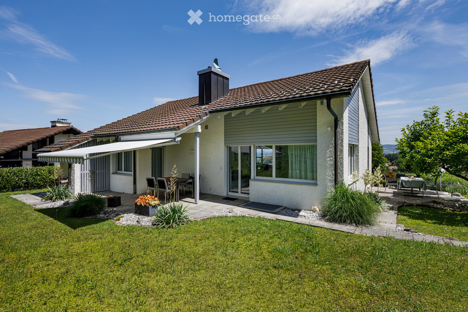 Single-story house with a tiled roof, white exterior, large windows, and a covered patio area. The front yard features a well-maintained lawn, garden beds, and outdoor seating.