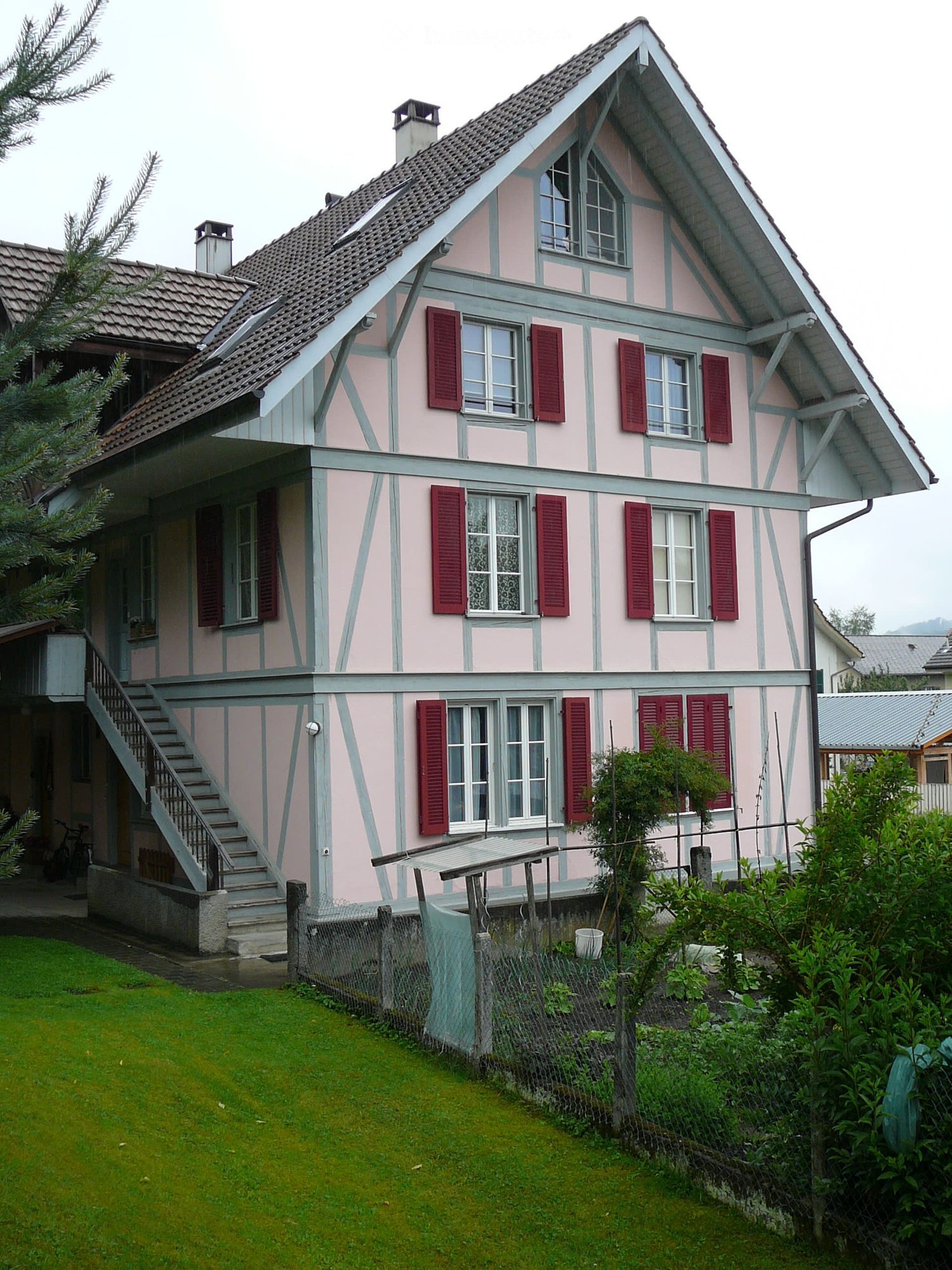 Two-story pink house with grey trim, red shutters, and a tiled roof. Features a staircase leading to the entrance and a small garden with a fence and plants.