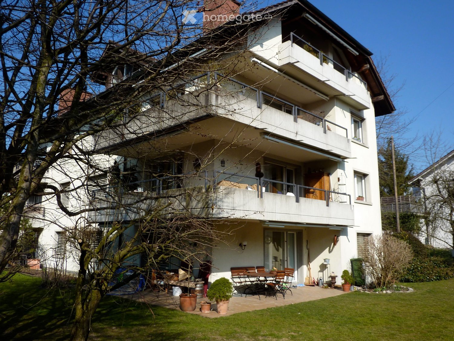 A two-story white house with balconies, surrounded by a well-maintained garden and patio area with outdoor furniture.