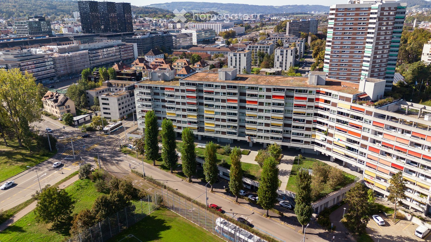 Aerial view of a modern apartment complex with colorful facades, surrounded by trees, roads, and other buildings in an urban setting.