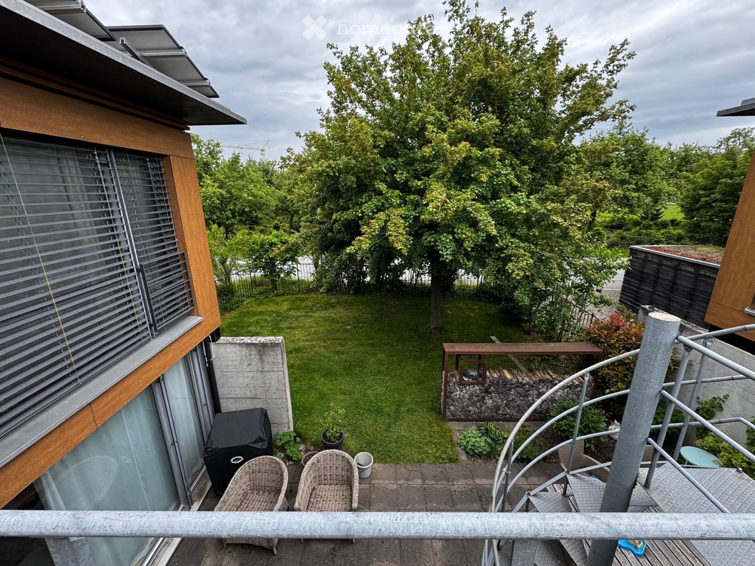 Balcony with two wicker chairs, a small table, and a view of a well-maintained garden with trees and a lawn.