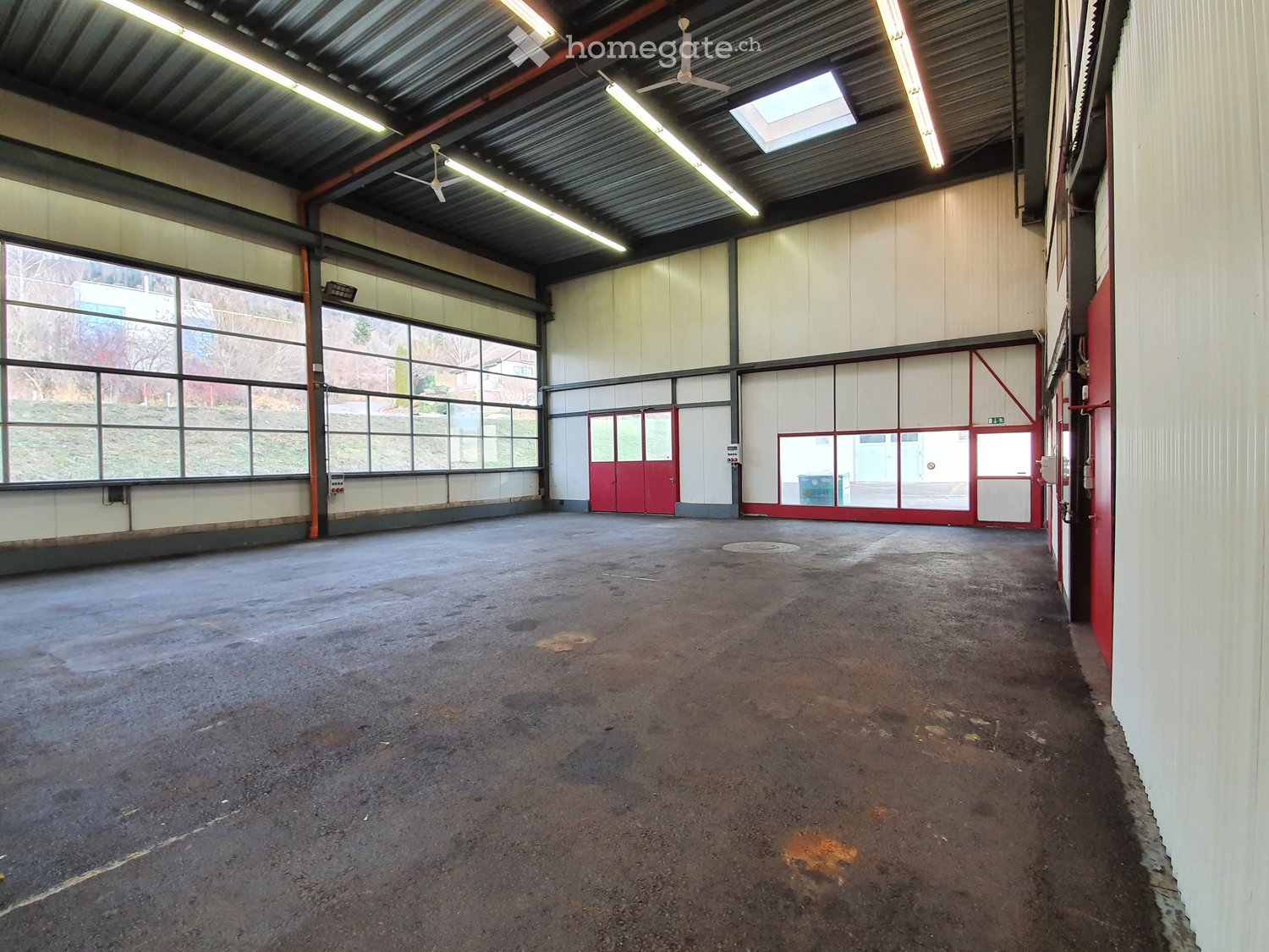 Large, empty industrial-style room with concrete floor, white walls, large windows, and red doors.