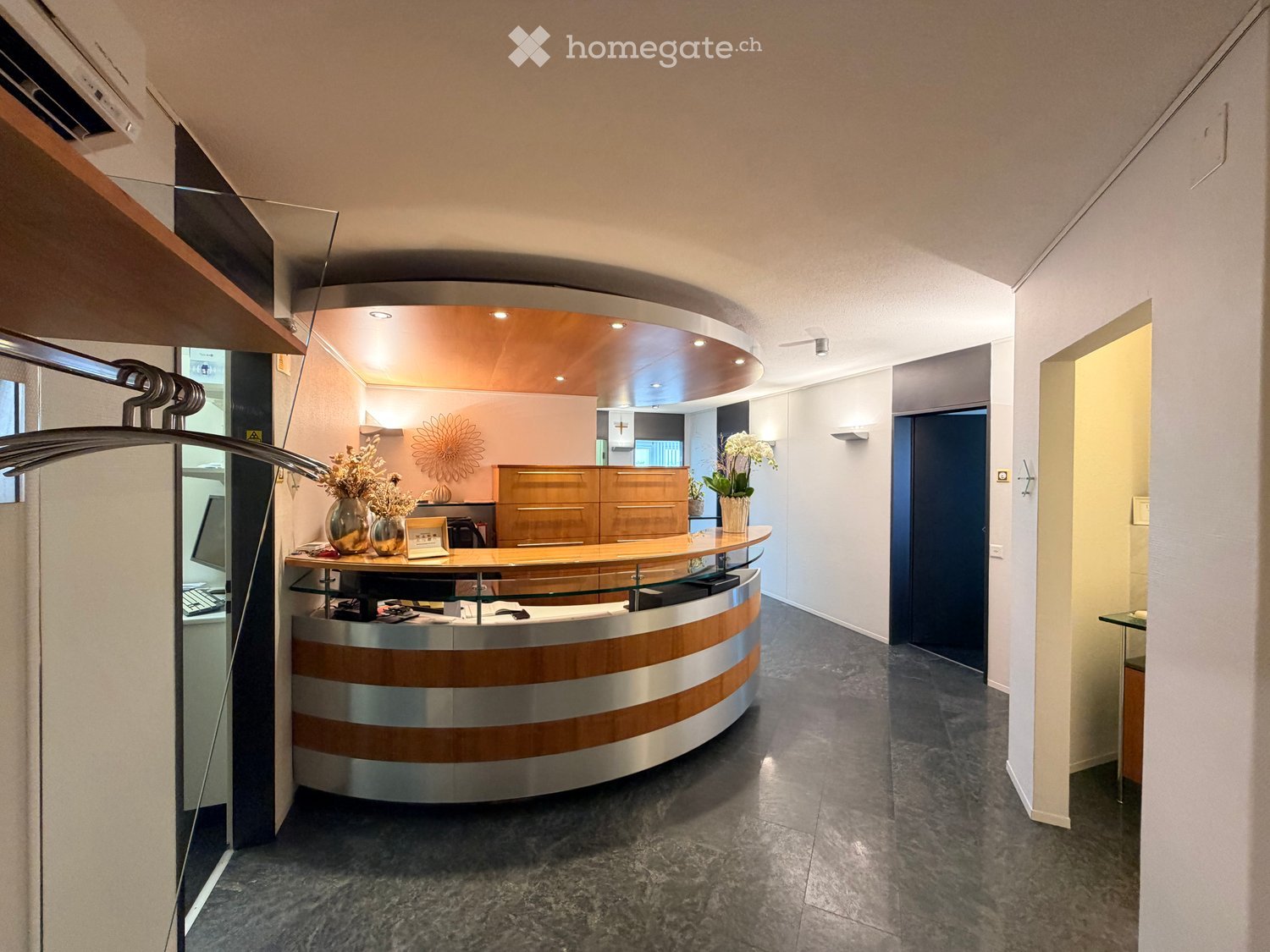 Modern reception area with a curved wooden and glass reception desk, vases with flowers, a screen, and a chair.