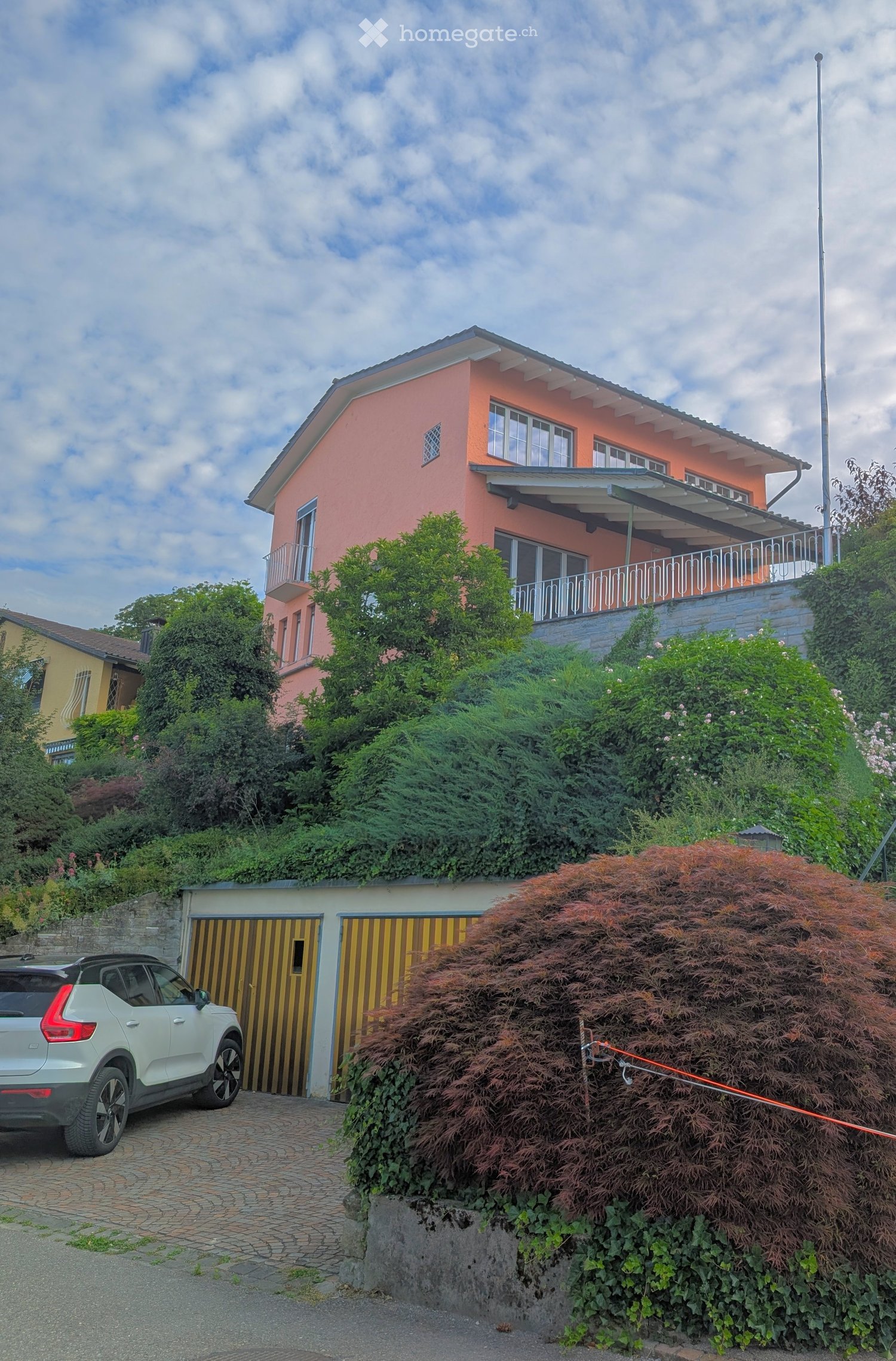 Two-story house with pink exterior, white-framed windows, and a balcony on the upper level. Garage with yellow striped doors is visible, along with a parked car.