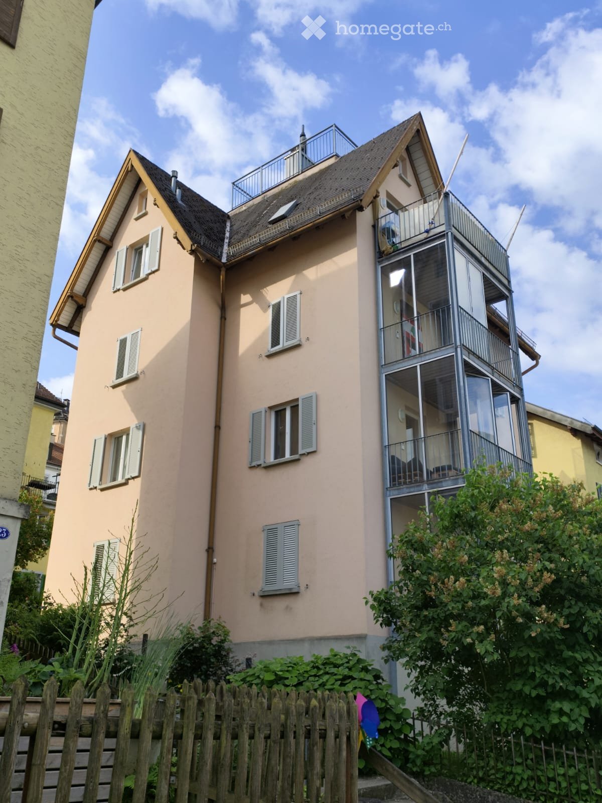Two-story house with beige facade, multiple windows with shutters, and a modern glass balcony on the upper level.
