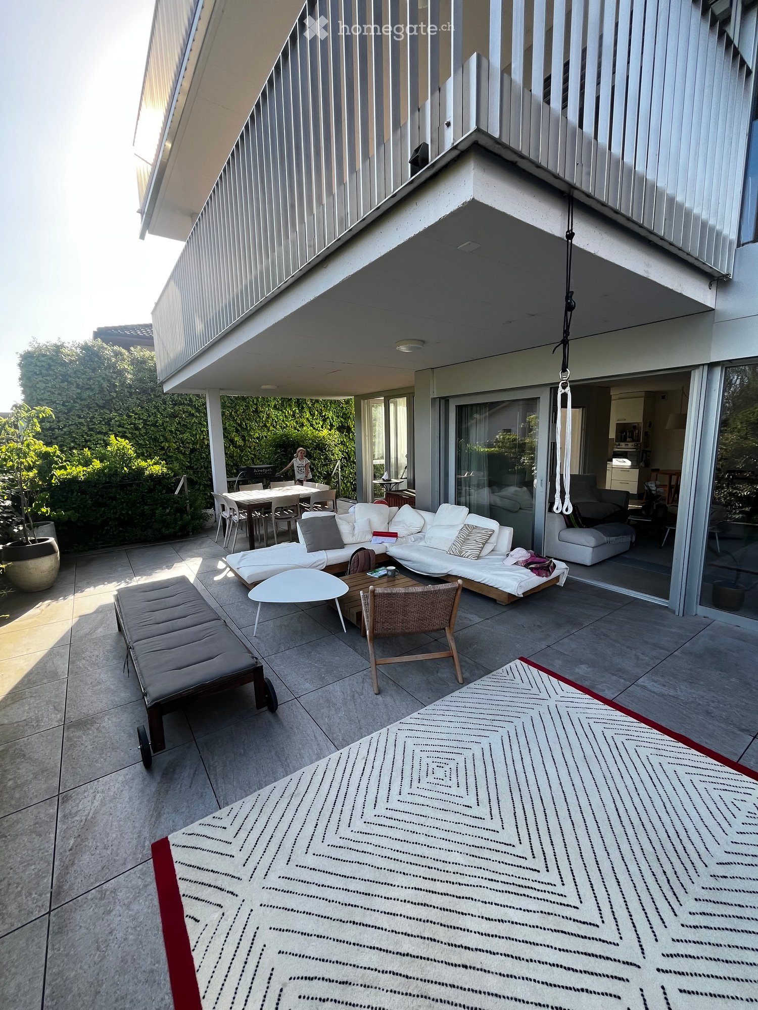 Modern outdoor terrace with a sofa, sun lounger, coffee table, chairs, and a patterned rug. The space is connected to the indoor living area through large glass doors.