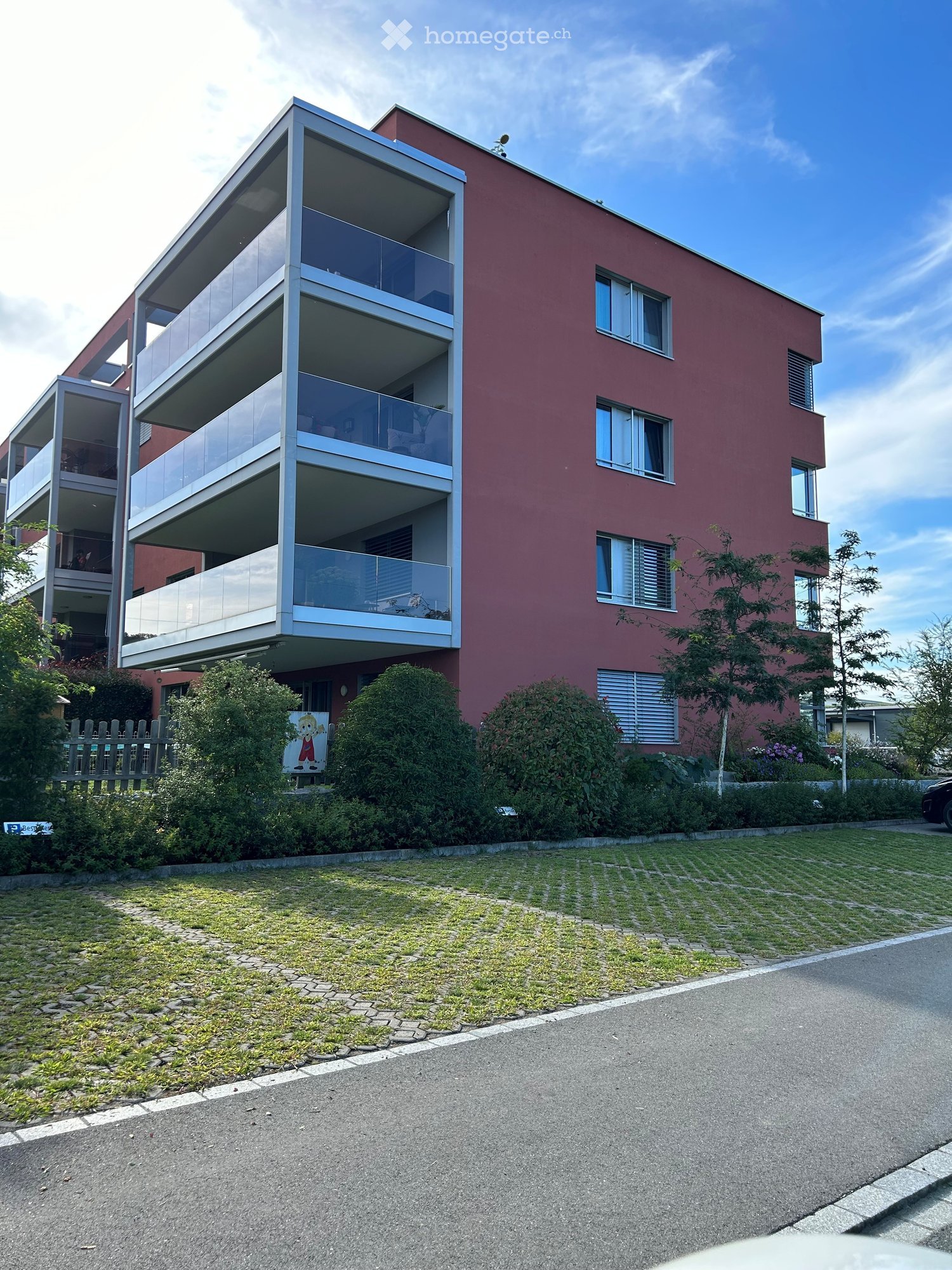 Modern apartment building, 3 stories, red exterior, large glass balconies, green lawn, sidewalk
