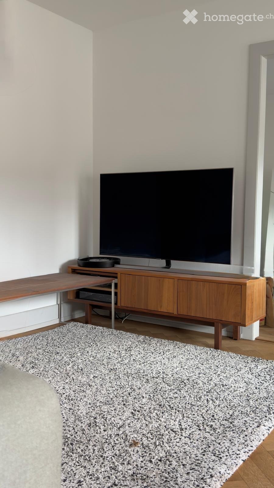Modern living room with a large flat-screen TV on a wooden cabinet, a wooden shelf, and a grey shaggy rug on the floor.