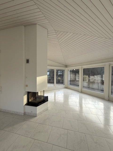 Bright, open room with a fireplace, large windows, and a sloped white ceiling. The space is completely empty with no furniture or equipment.