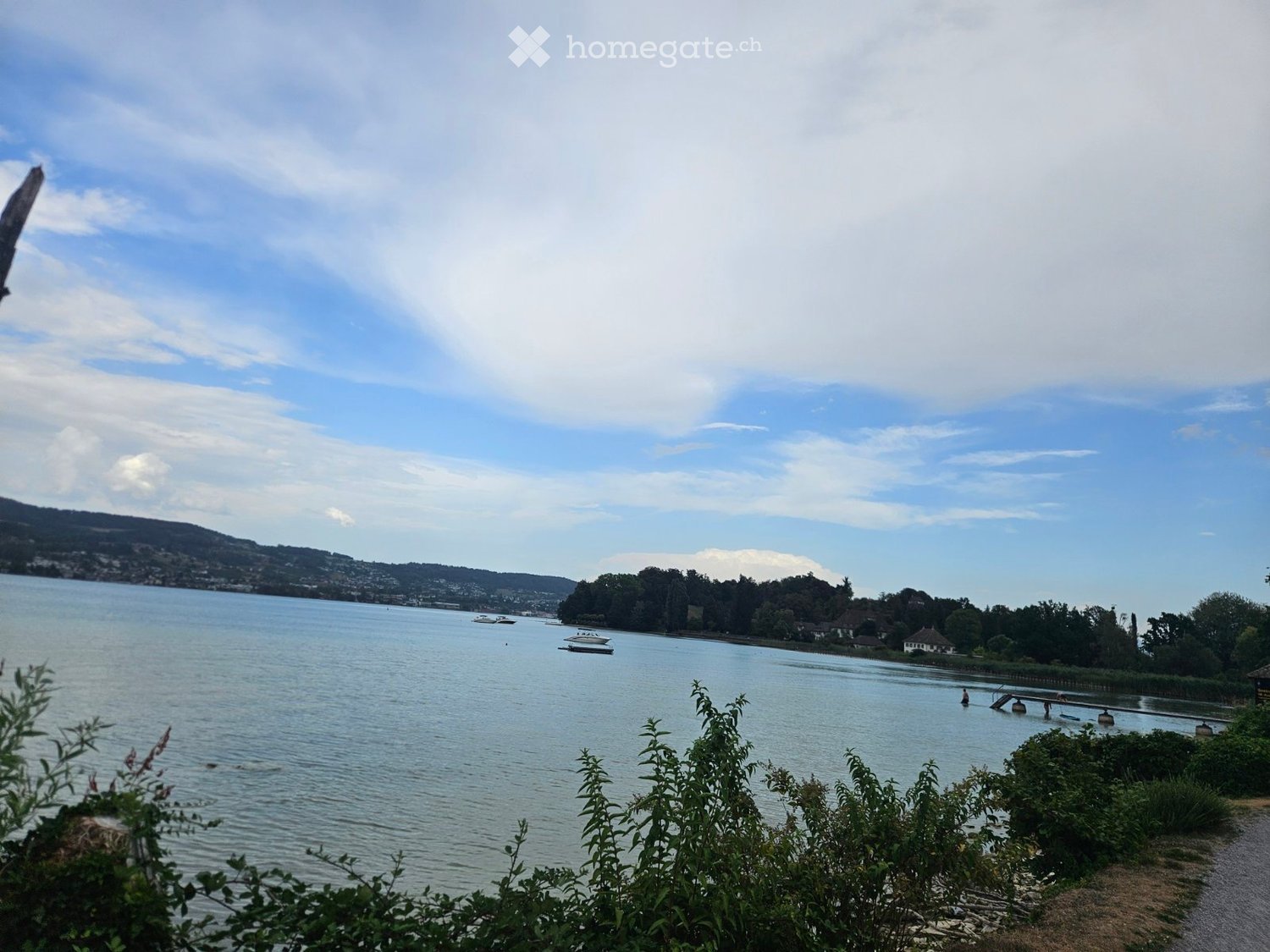 Scenic view of a lake with boats, surrounded by trees and hills under a partly cloudy sky.