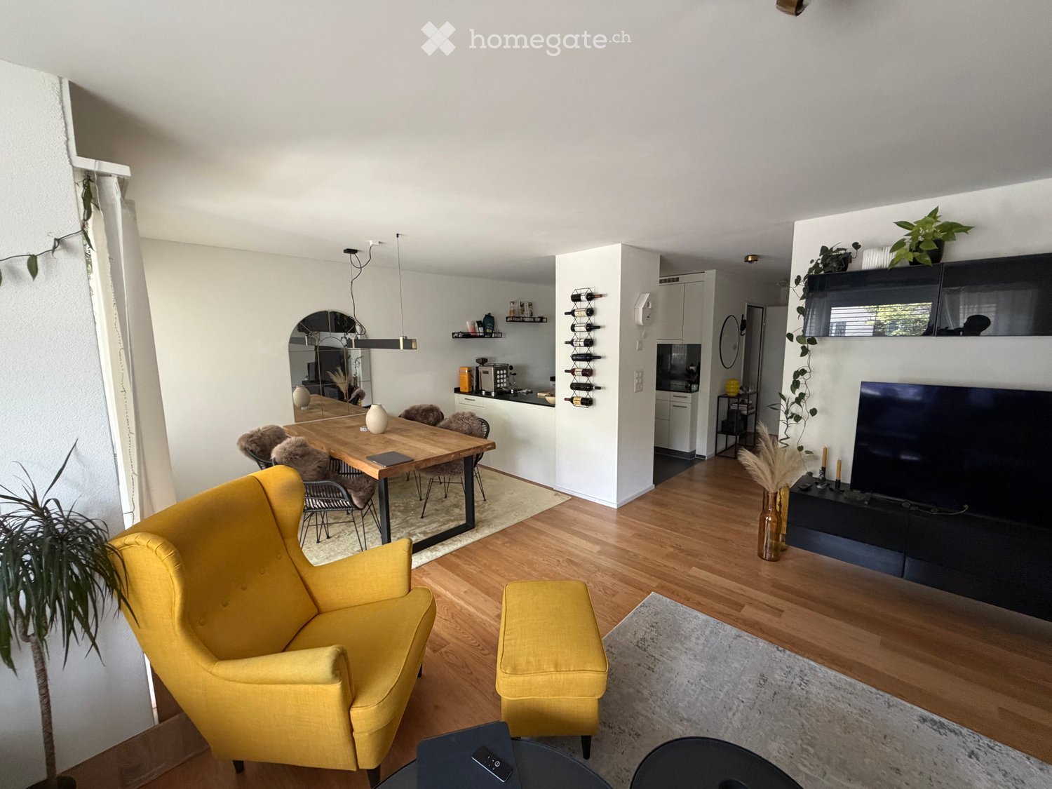 Bright living and dining area with wooden floors, yellow sofa, small round table, dining table with chairs, TV, wine rack, and kitchen area visible.