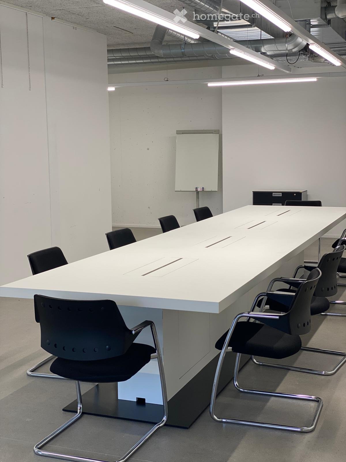 long desk, several black chairs, white wall, ceiling lamp