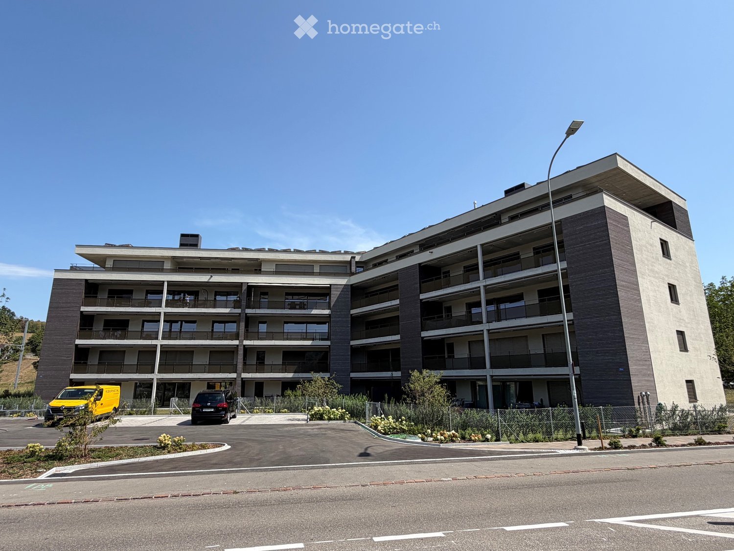 Modern multi-story residential building with balconies, large windows, and a contemporary design. The building features a mix of light and dark tones, with a parking area and landscaped surroundings.