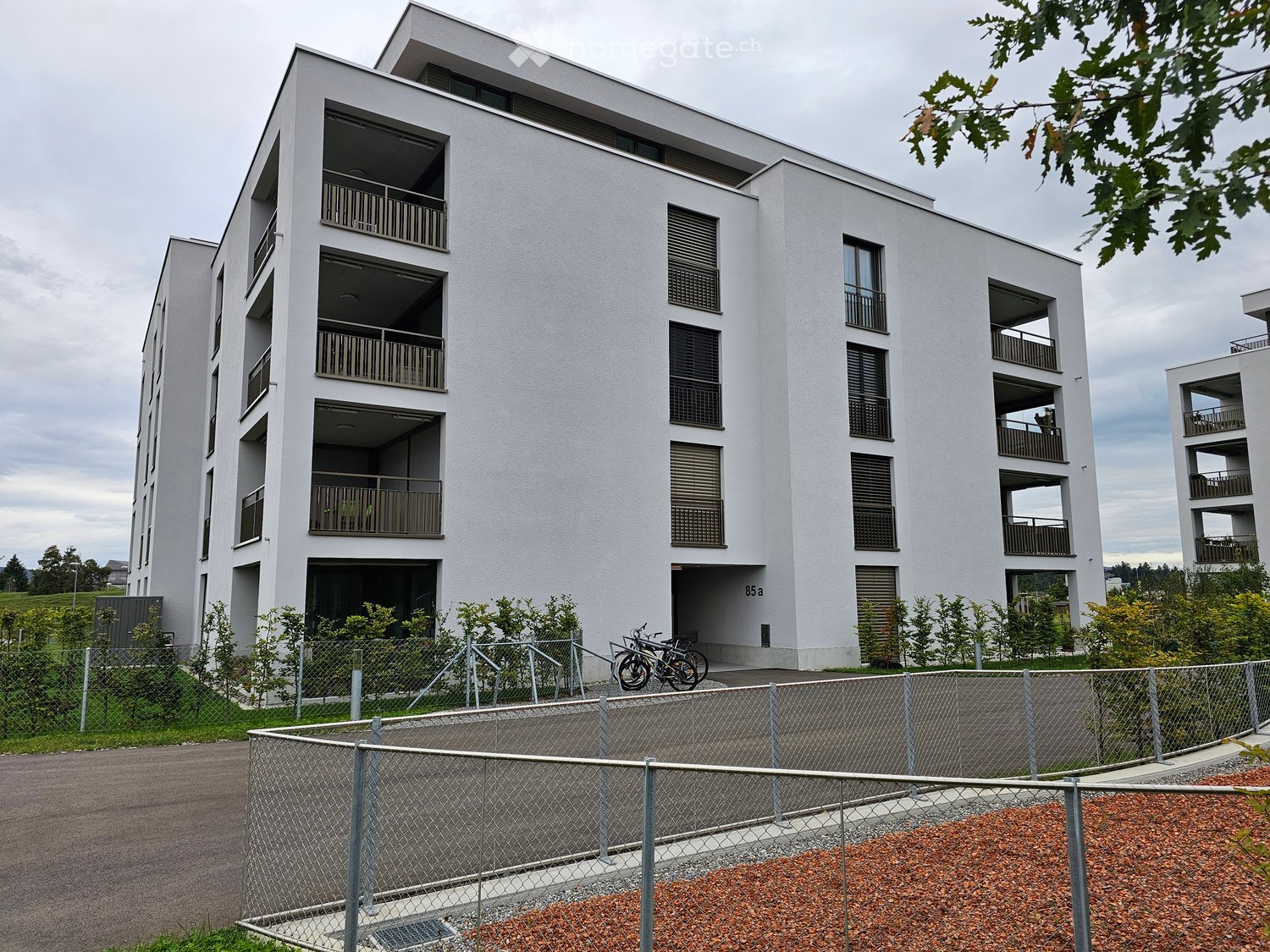 Modern multi-story residential building with white facade, multiple balconies, and a fenced area in front.