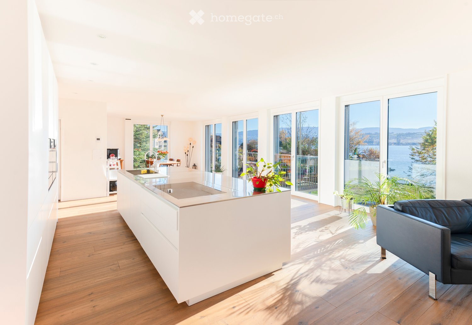Bright, modern kitchen with white cabinetry, a central island, and large windows offering a scenic view. Includes a black leather sofa, potted plants, and a dining area with a table and chairs.