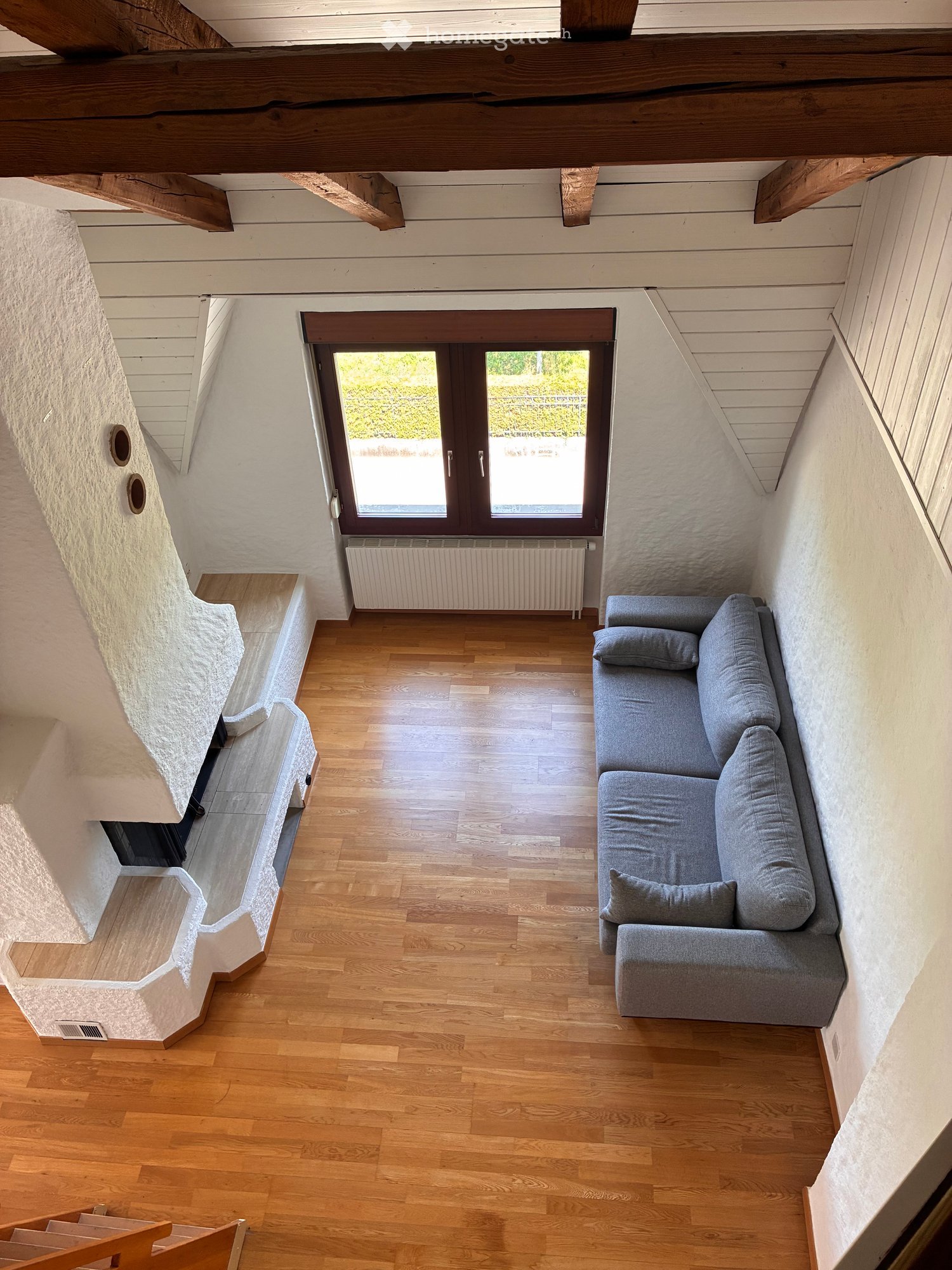Cozy living room with wooden flooring, exposed wooden beams, a grey sectional sofa, and a radiator beneath a large window.